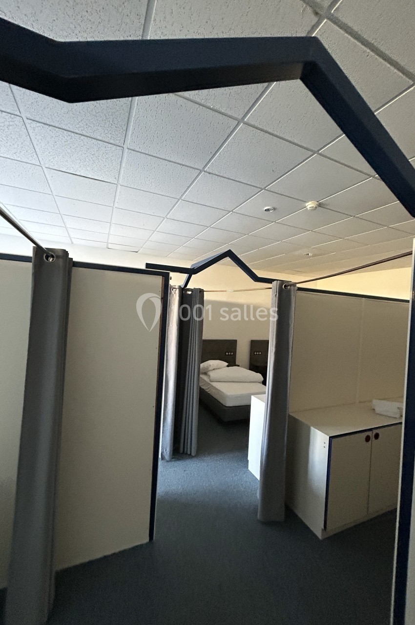 Cloisons modulaires délimitant des espaces avec lits simples, rideaux et mobilier dans une pièce éclairée.