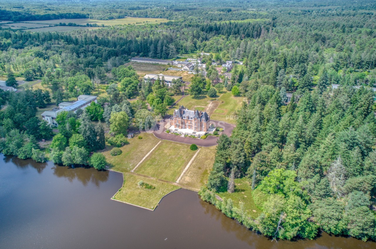 Vue aérienne d'un manoir entouré de forêt, avec un lac au premier plan et des bâtiments en arrière-plan.