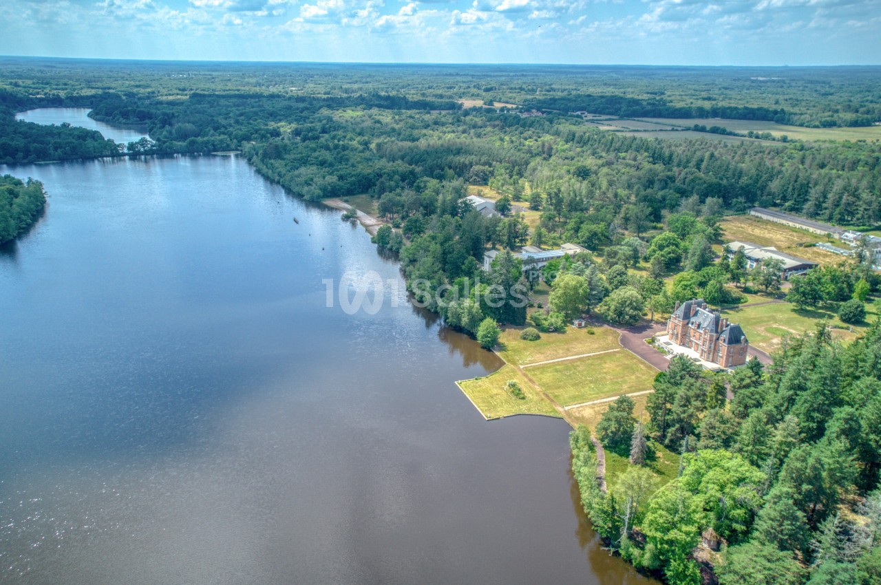 Vue aérienne d'un lac entouré de forêts et de prairies, avec un bâtiment en bordure et un ciel dégagé.