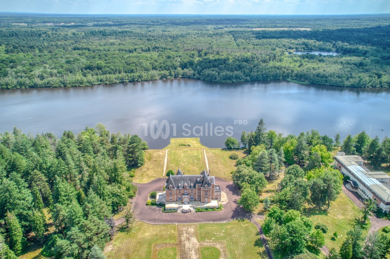 Vue aérienne d'un château entouré de forêt, situé au bord d'un grand lac sous un ciel dégagé.