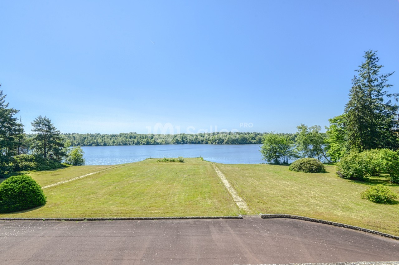 Vue sur un grand jardin verdoyant menant à un lac entouré d'arbres sous un ciel bleu dégagé.