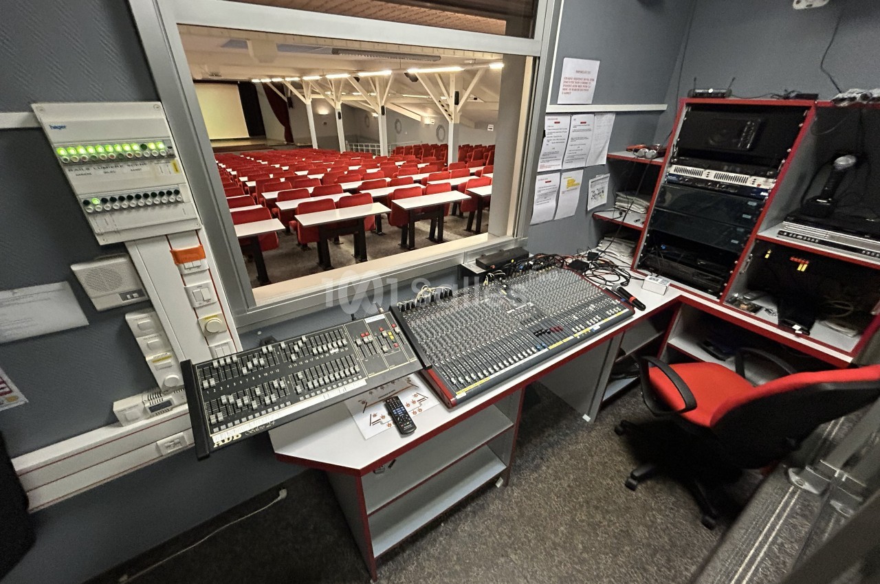 Salle de régie équipée de consoles de mixage audio et vidéo, avec vue sur une salle de conférence vide.