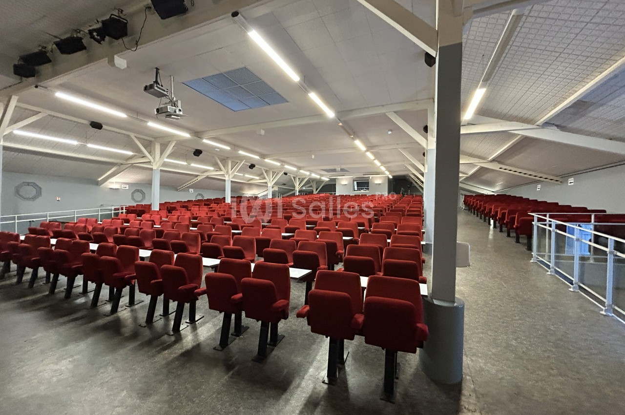 Salle de conférence vide avec des rangées de sièges rouges, éclairée par des lumières au plafond.