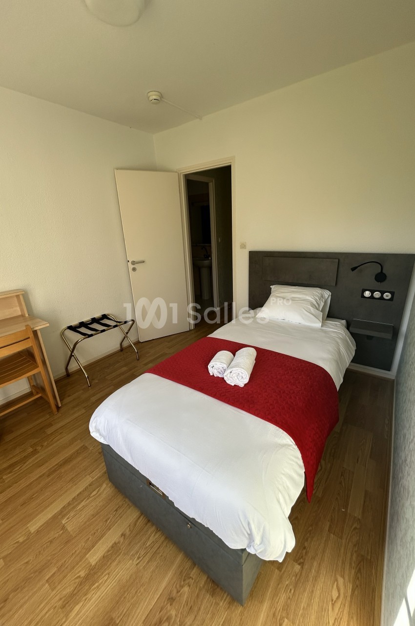 Chambre simple avec lit une place, drap rouge, bureau en bois, porte ouverte sur une autre pièce.