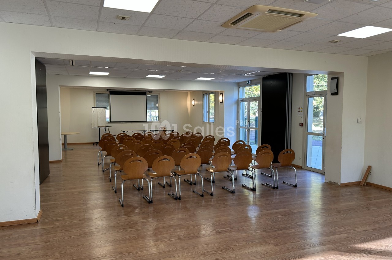 Salle de conférence lumineuse avec des rangées de chaises en bois, un écran de projection et un tableau blanc.