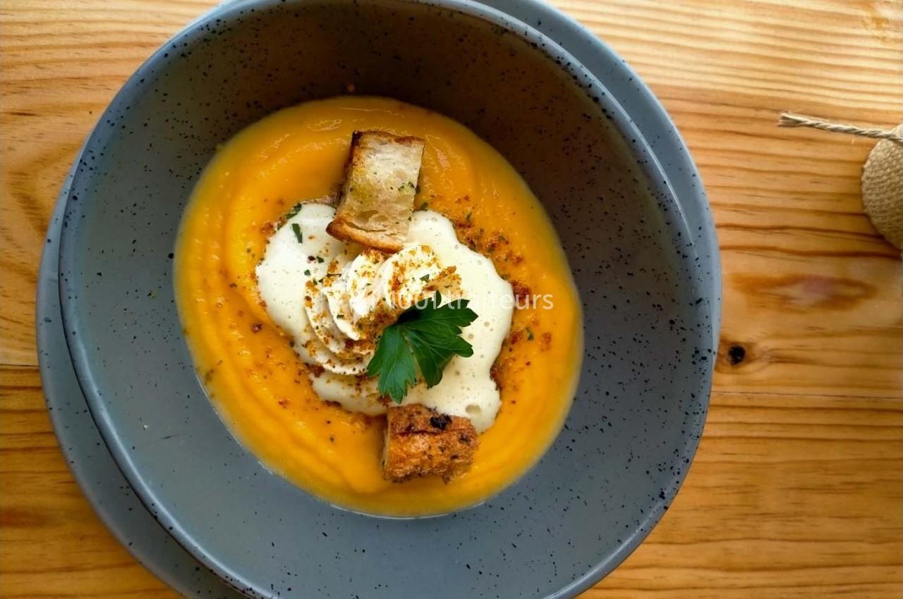 Bol de soupe de potiron garnie de croûtons, crème fraîche, persil et épices, posé sur une table en bois.