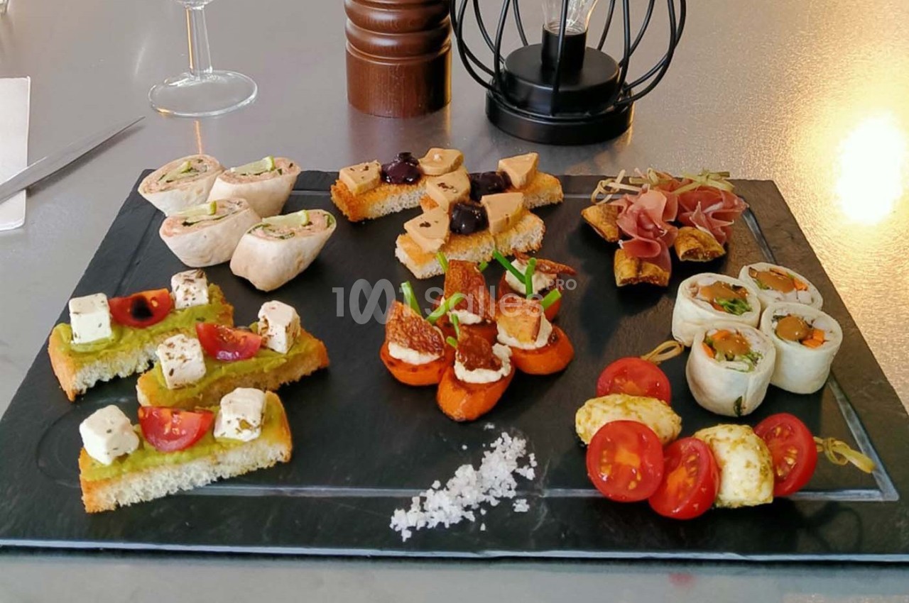 Plateau d'amuse-bouches variés comprenant toasts, roulés, légumes, fromage et charcuterie, présenté sur une ardoise.