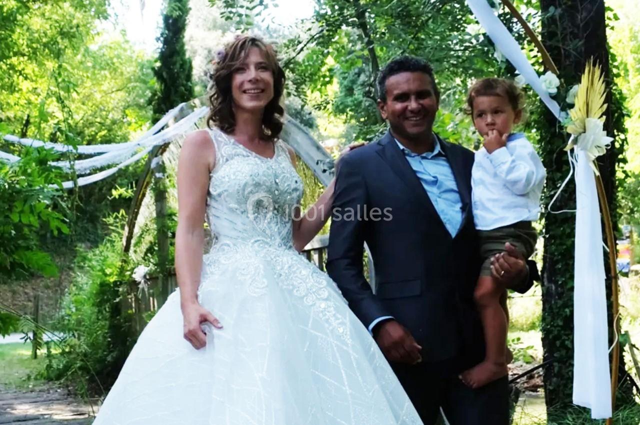 Un couple en tenue de mariage pose avec un enfant dans un jardin décoré de rubans et de fleurs.