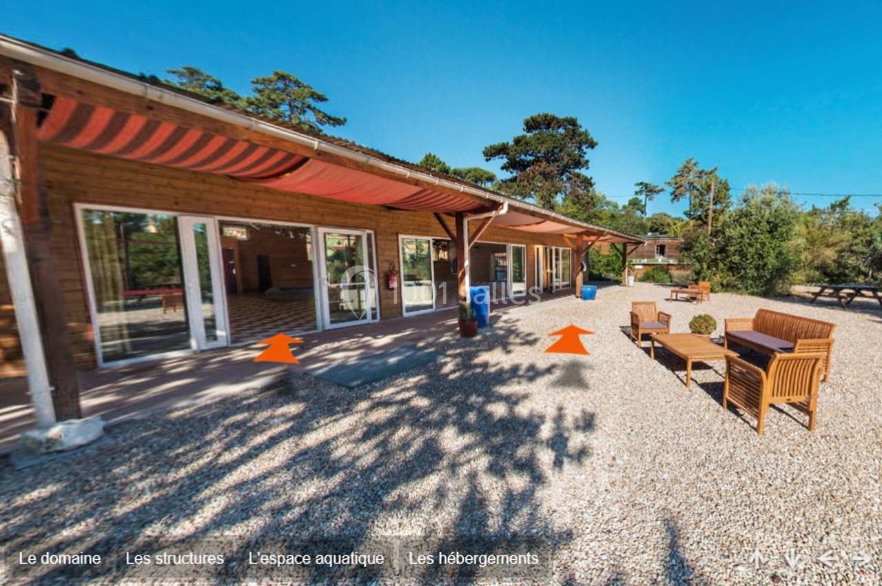 Terrasse en gravier avec mobilier en bois, devant un bâtiment en bois aux larges baies vitrées et stores rouges.