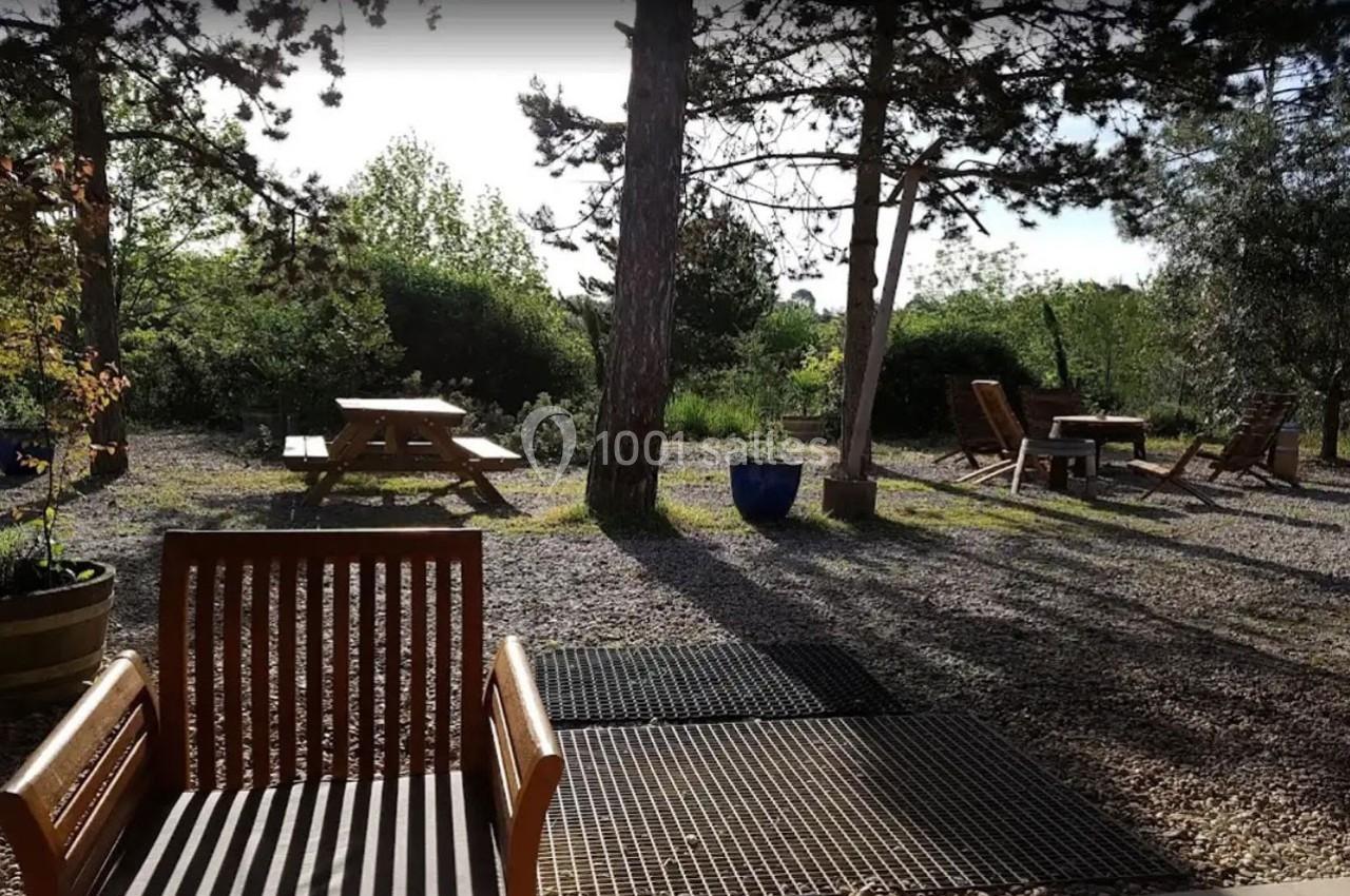 Chaises en bois et tables de pique-nique disposées dans un jardin ombragé entouré de végétation.