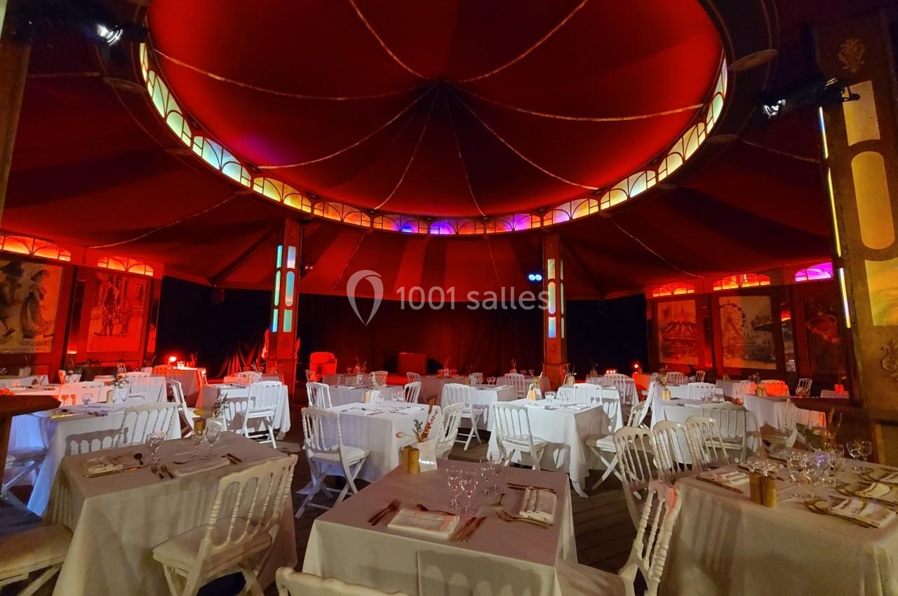 Intérieur d'un chapiteau avec tables dressées, chaises blanches et éclairage chaleureux rouge et jaune.
