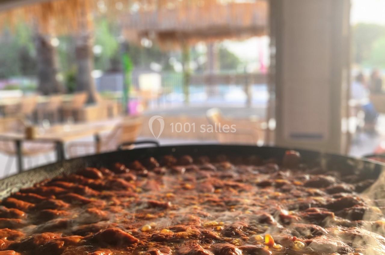 Grande poêle de paella en cuisson avec des morceaux de viande, en extérieur sous une terrasse ombragée.