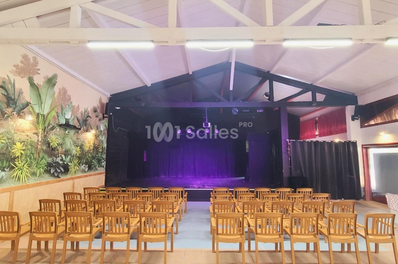 Salle avec des rangées de chaises en bois face à une scène éclairée par des projecteurs violets, décor mural végétal.