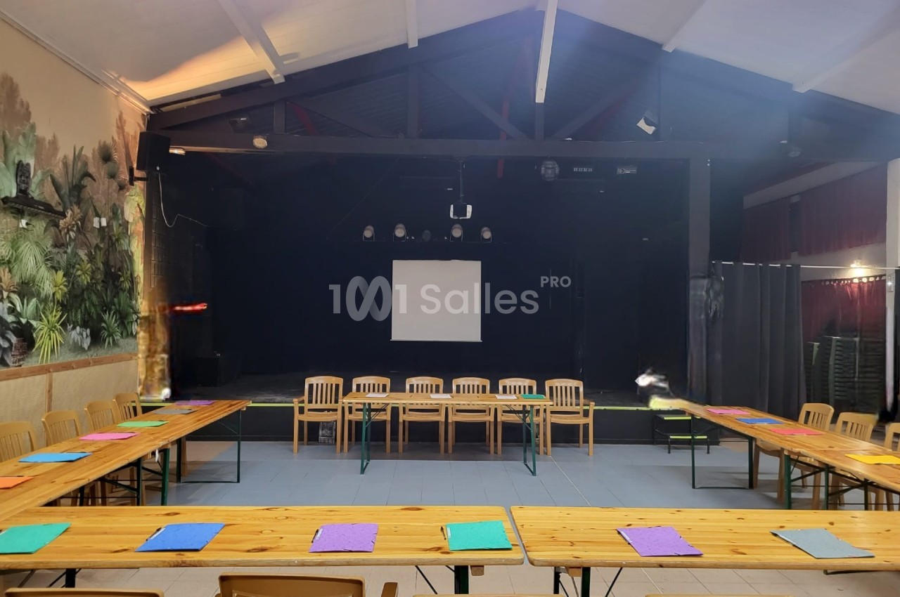 Salle avec une scène, des tables en U disposées avec des feuilles colorées et des chaises en bois, éclairage intérieur.