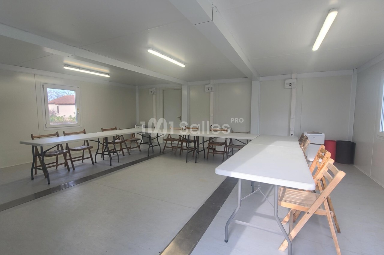 Salle lumineuse avec tables blanches disposées en U, chaises en bois et murs clairs dans un espace modulaire.