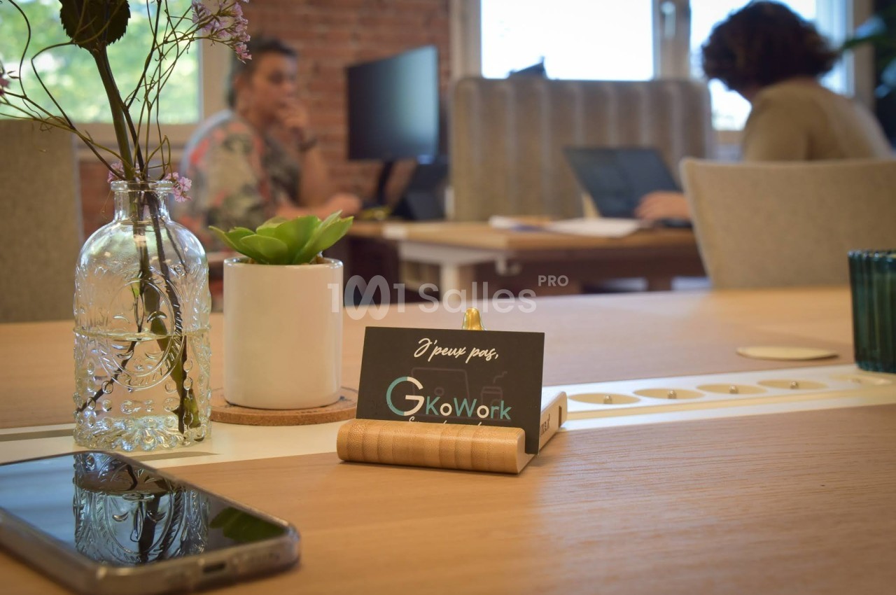 Bureau partagé avec plantes, carte posée sur un support en bois, et personnes travaillant en arrière-plan.