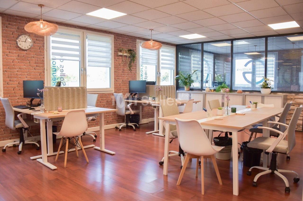 Espace de travail lumineux avec bureaux partagés, chaises modernes, plantes et murs en briques apparentes.