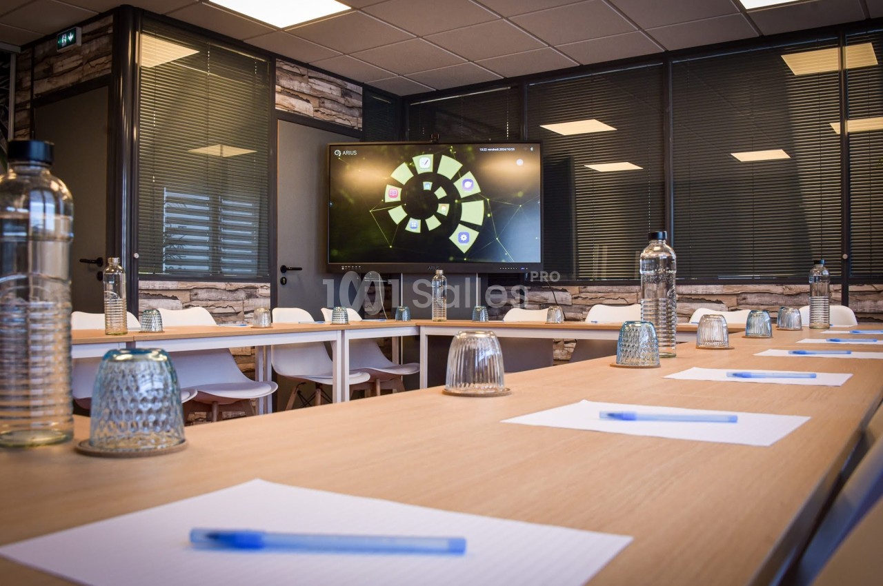 Salle de réunion moderne avec table en U, chaises blanches, bouteilles d'eau, blocs-notes, stylos et écran allumé.