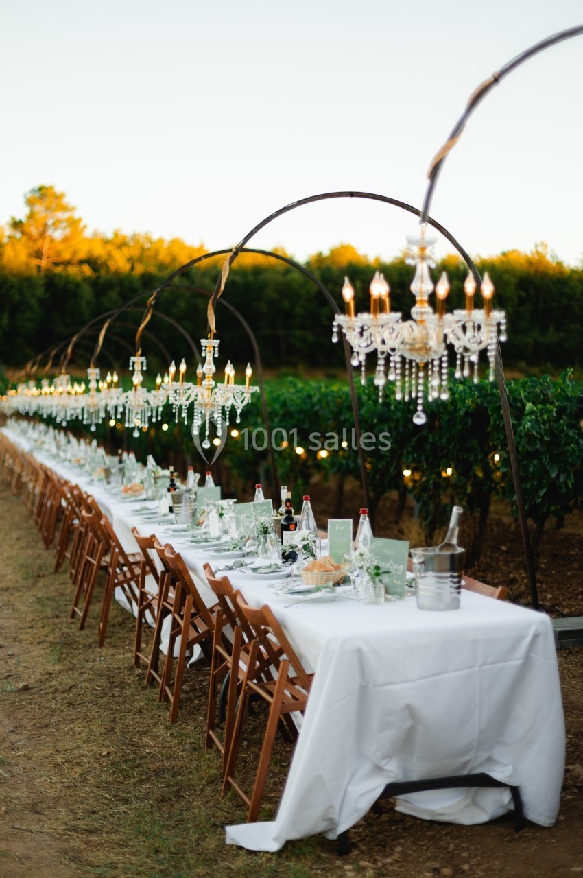 Grande table dressée en extérieur avec nappes blanches, chaises en bois et lustres suspendus, entourée de vignes.