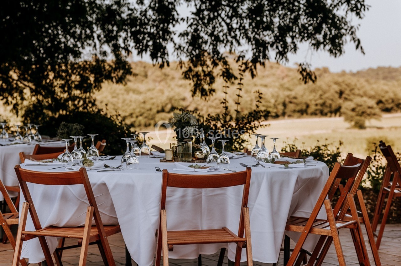 Table dressée en extérieur avec nappes blanches, chaises en bois et verres, entourée de verdure et d'un paysage champêtre.