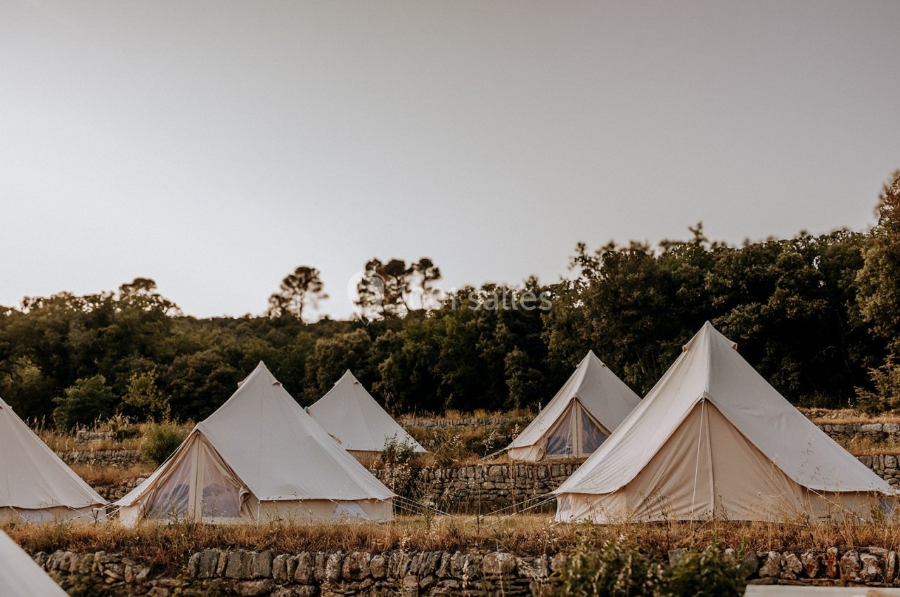 Tentes en toile beige installées sur un terrain herbeux entouré d'arbres et de murets en pierre.