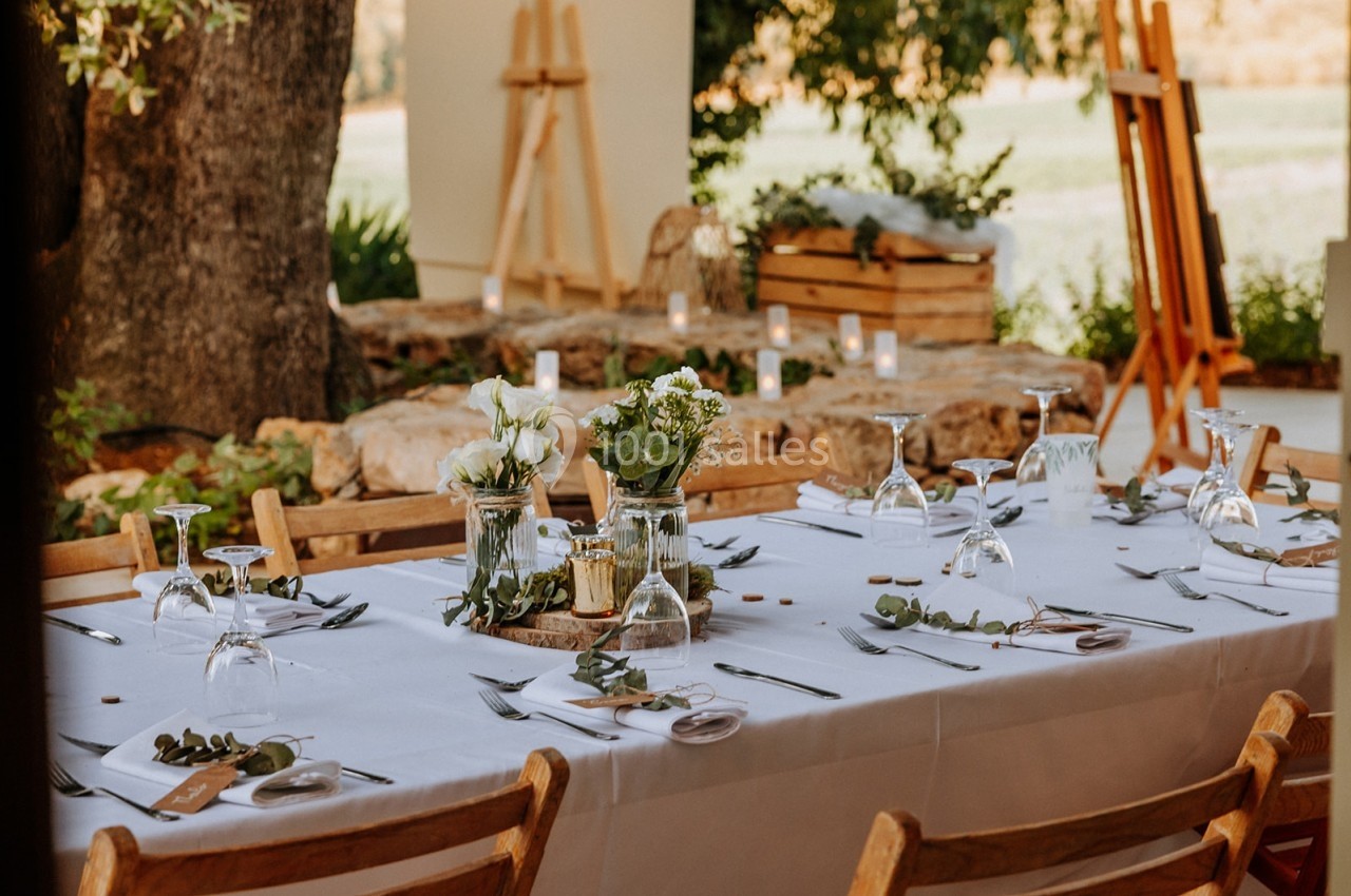 Table décorée pour un repas en plein air avec nappes blanches, verres, couverts et arrangements floraux.