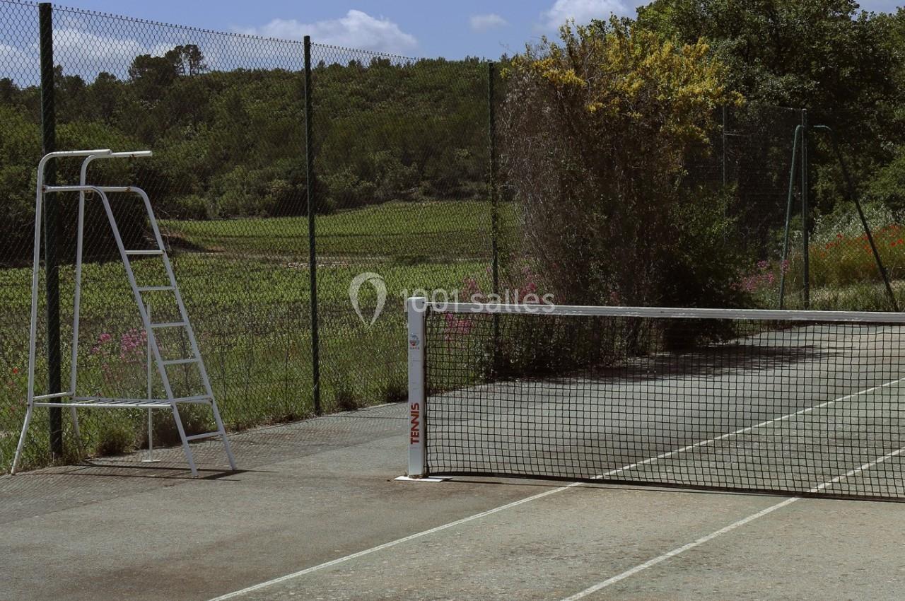 Terrain de tennis extérieur avec filet, chaise d'arbitre et clôture, entouré de végétation et d'arbres.