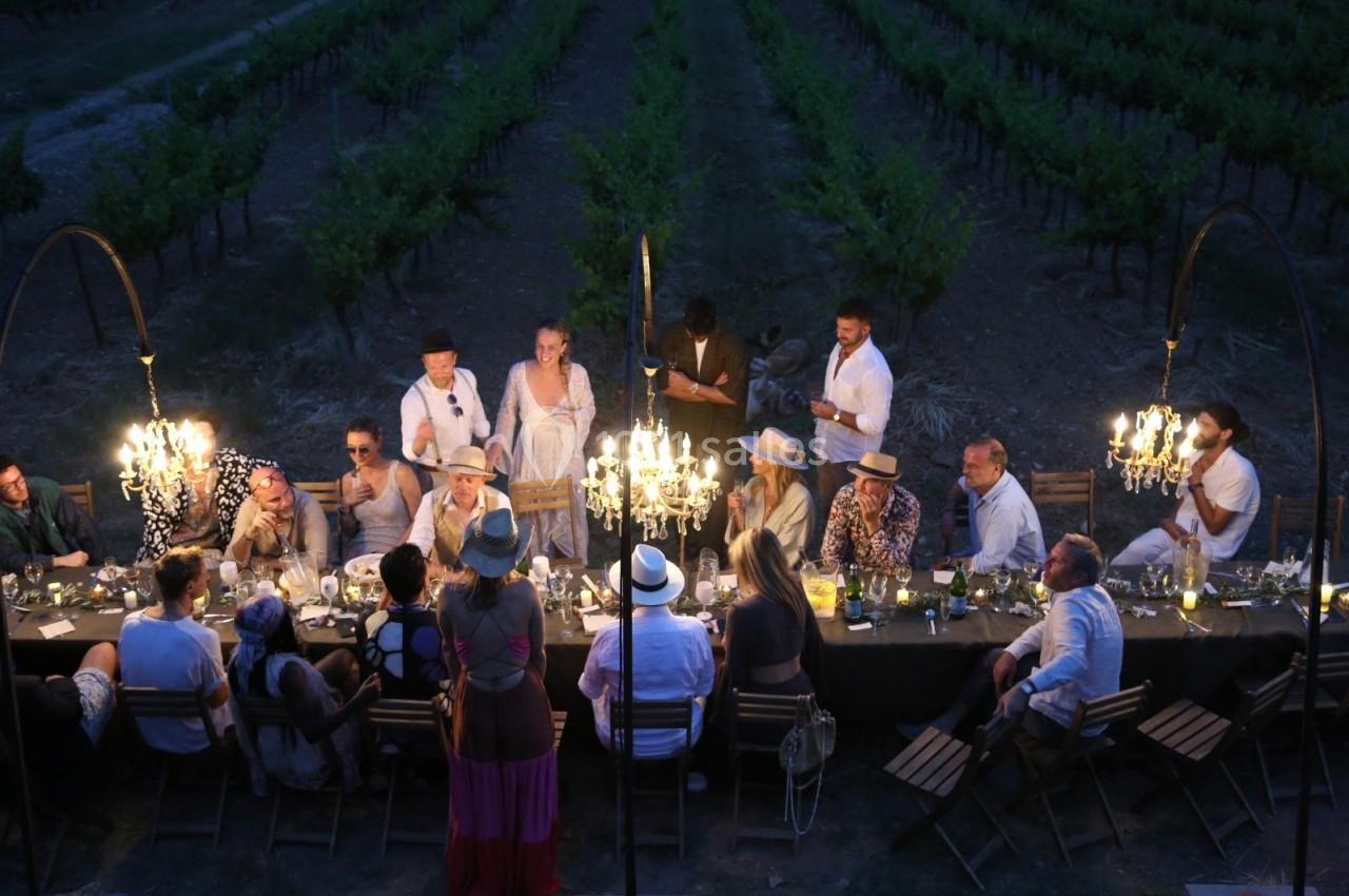 Groupe de personnes dînant en plein air autour d'une longue table éclairée par des lustres, près de vignes.