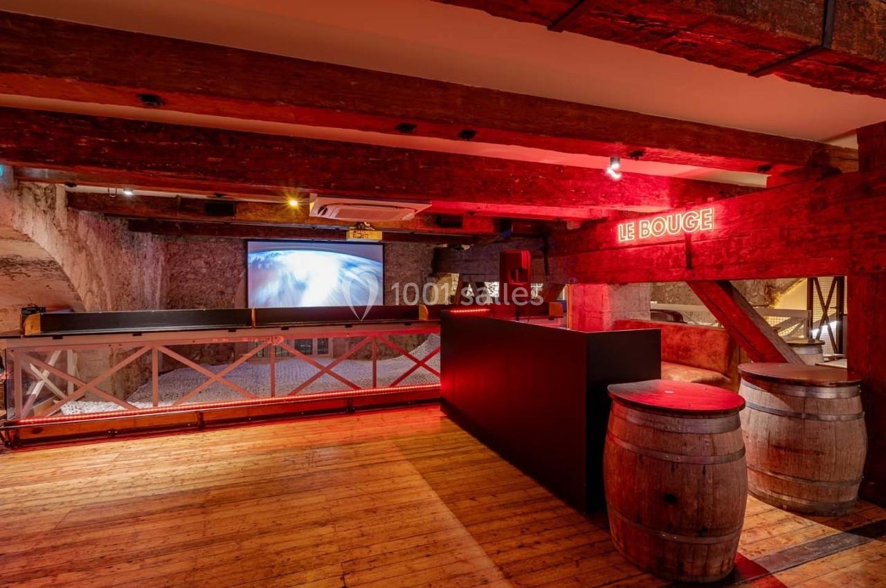 Salle avec poutres apparentes, bar en bois, tonneaux décoratifs, écran mural et enseigne lumineuse rouge.