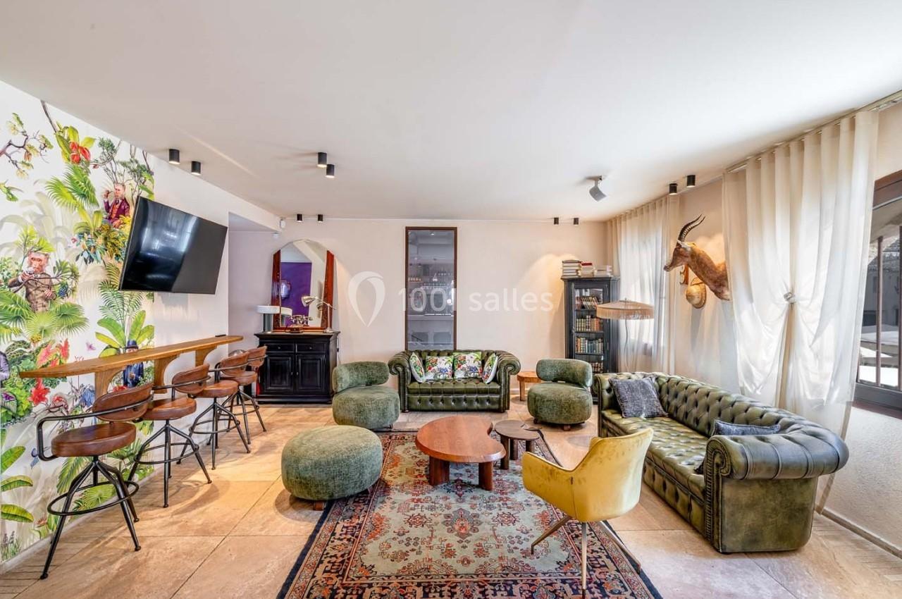 Salon lumineux avec canapés verts, fauteuils, table basse, bar avec tabourets, murs décorés et grande fenêtre avec rideaux.
