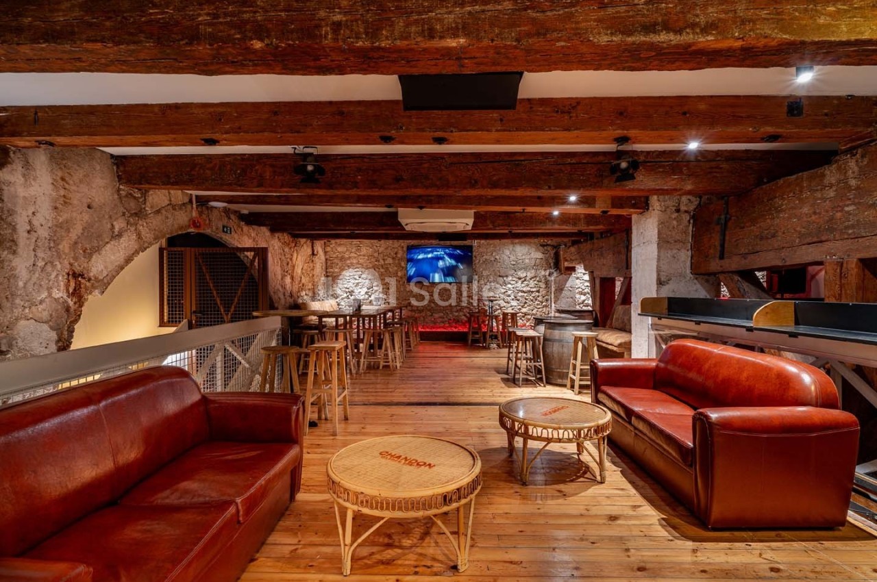 Salle avec murs en pierre, poutres apparentes, canapés en cuir rouge, tables en bois et écran mural allumé.