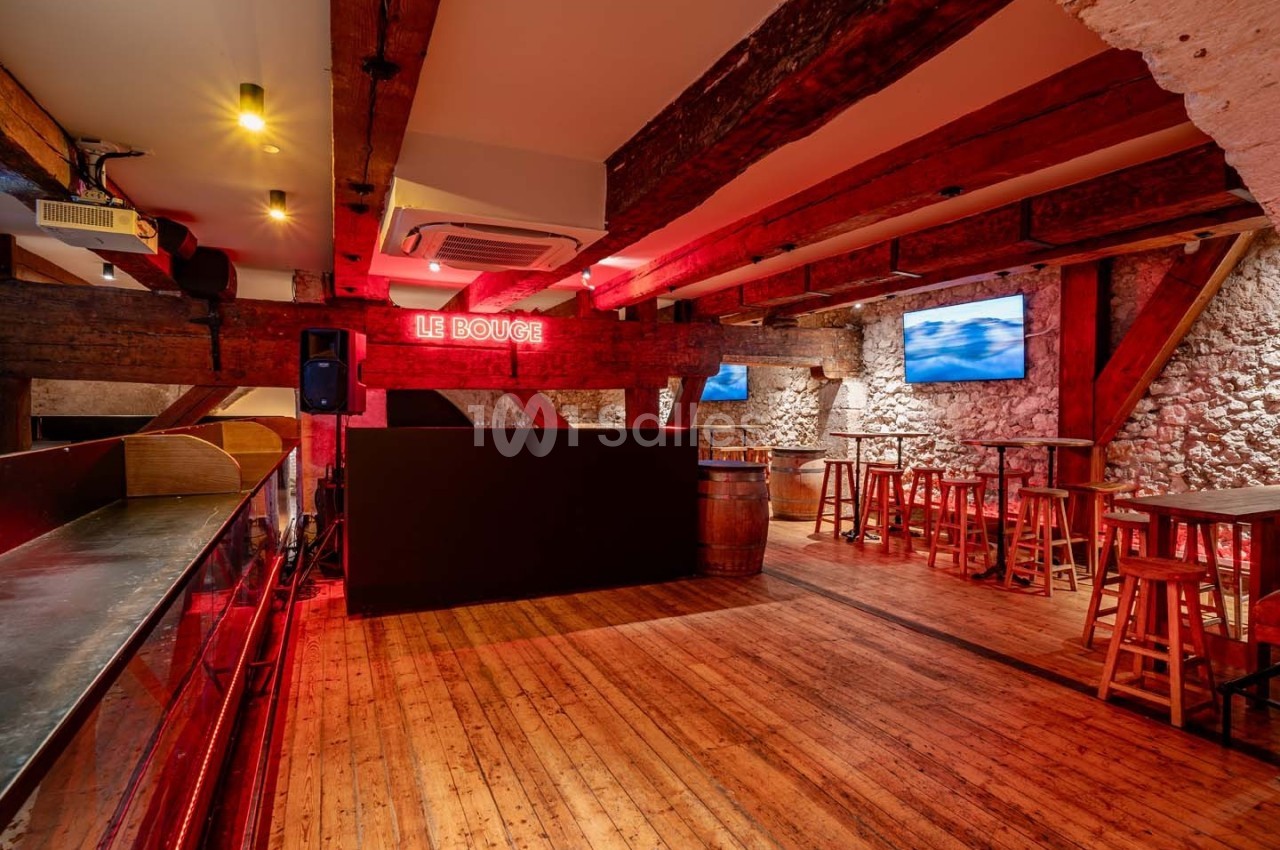 Salle intérieure avec poutres en bois apparentes, murs en pierre, bar à gauche et tables hautes avec tabourets.