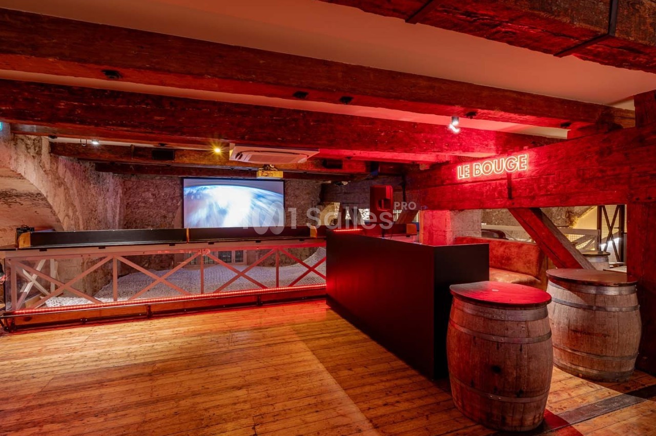 Salle avec poutres en bois apparentes, bar noir, tonneaux en décoration et écran projetant une image lumineuse.