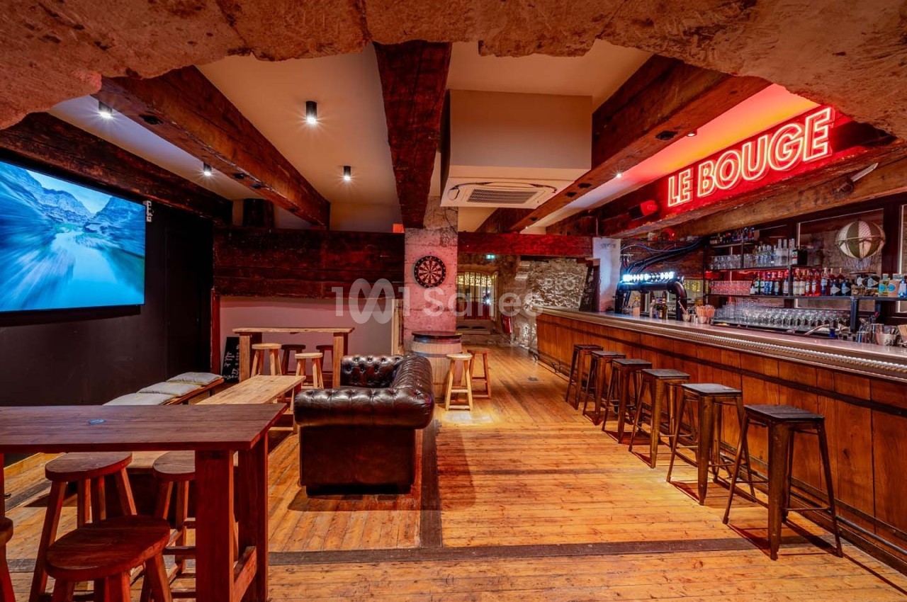 Intérieur d'un bar avec comptoir en bois, tabourets alignés, tables, canapé en cuir et enseigne lumineuse ’Le Bouge’.