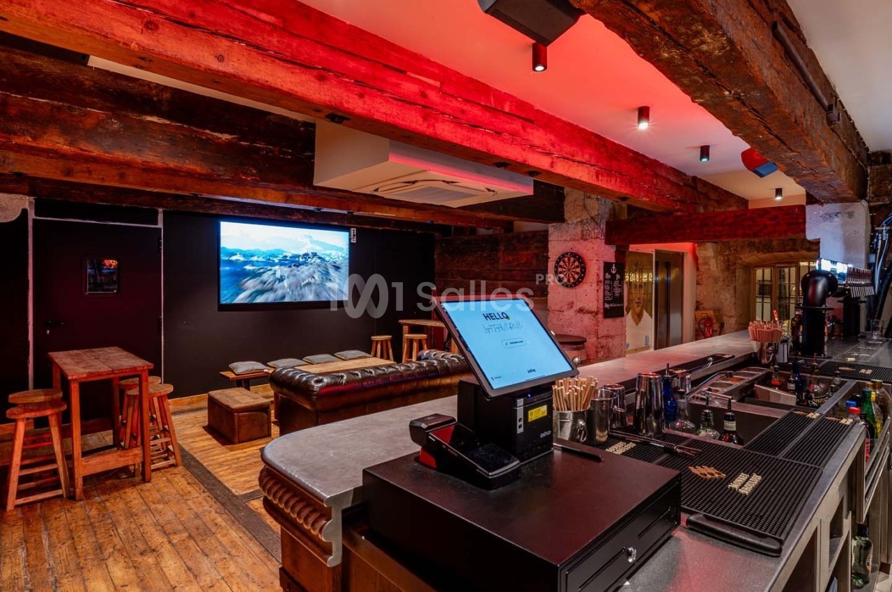 Intérieur d'un bar avec comptoir, tabourets, canapés en cuir, écran mural et éclairage chaleureux.