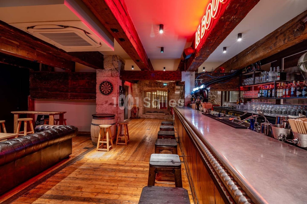 Intérieur d'un bar avec comptoir en bois, tabourets, canapé en cuir, fléchettes au mur et éclairage néon rouge.