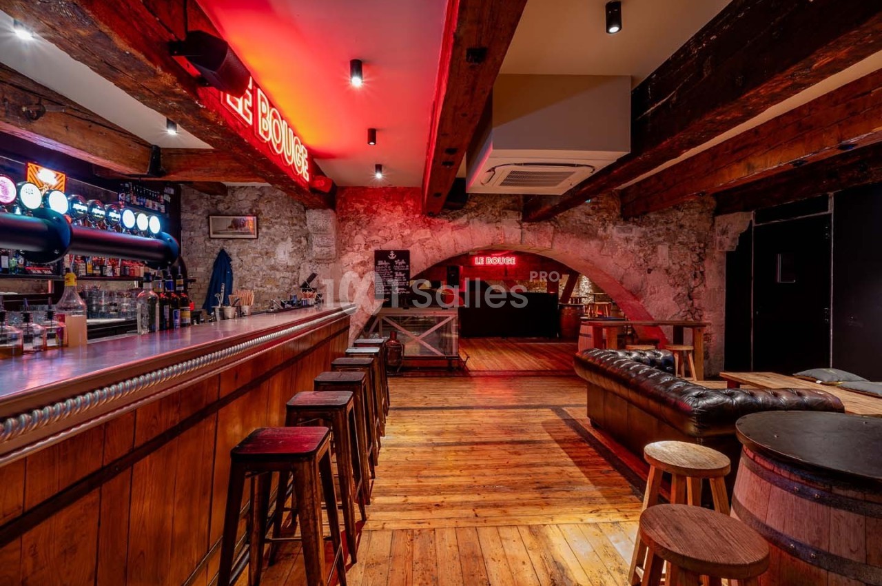 Intérieur d'un bar avec comptoir en bois, tabourets, canapés en cuir, éclairage rouge et murs en pierre apparente.