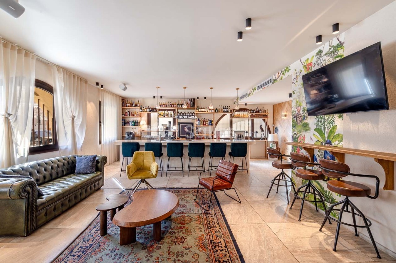Salon moderne avec canapé en cuir, bar équipé, tabourets hauts, tapis coloré et décoration murale végétale.