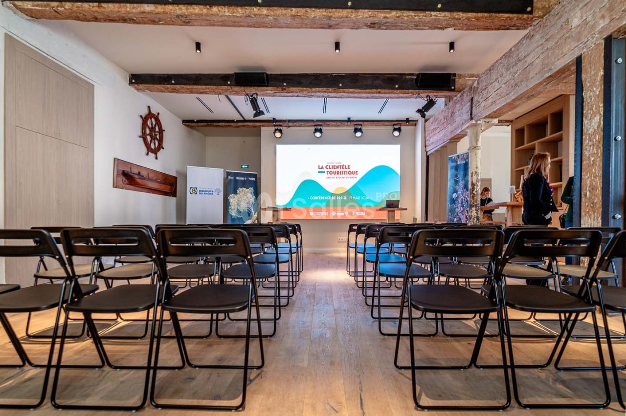 Salle de conférence avec des rangées de chaises noires face à un écran de projection allumé.