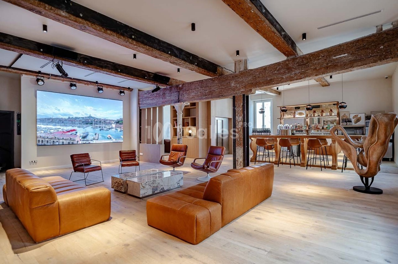 Salon spacieux avec canapés en cuir, bar en bois, poutres apparentes et écran mural affichant un paysage côtier.