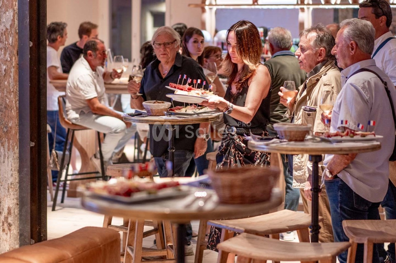 Des personnes discutent et dégustent des amuse-bouches dans un espace convivial avec des tables hautes en bois.