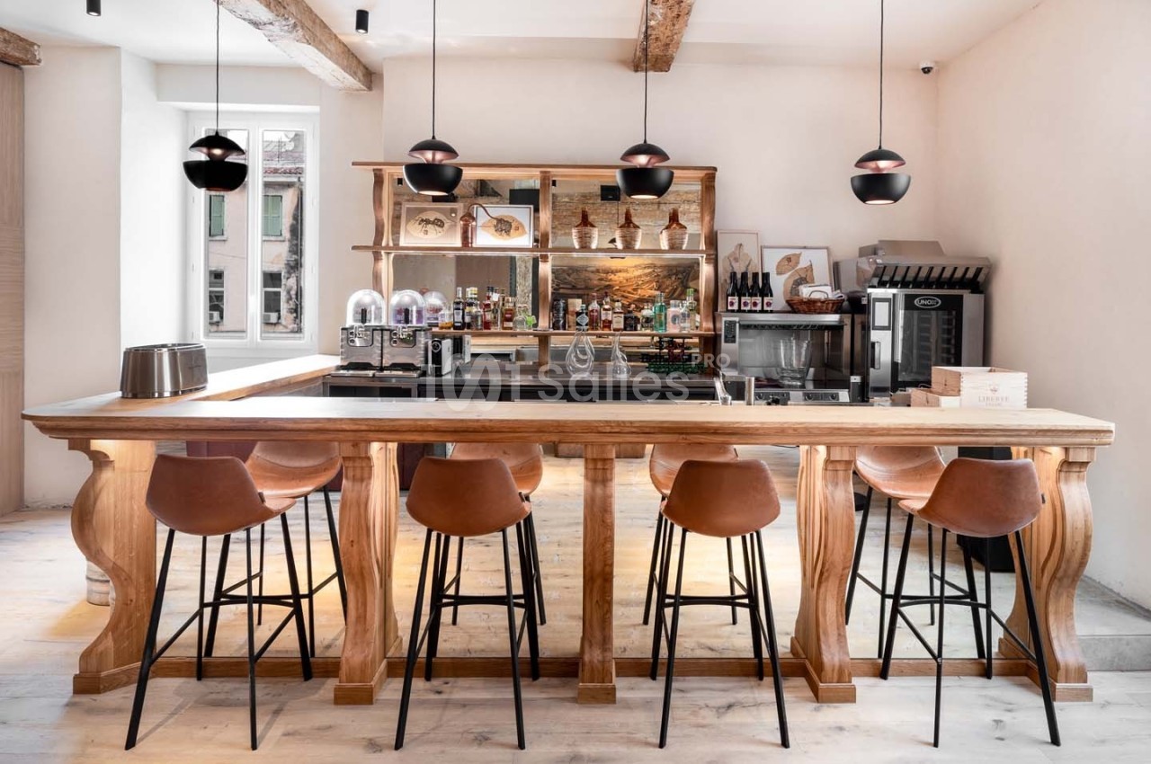 Bar en bois clair avec tabourets en cuir, étagères garnies de bouteilles et luminaires suspendus dans un intérieur lumineux.