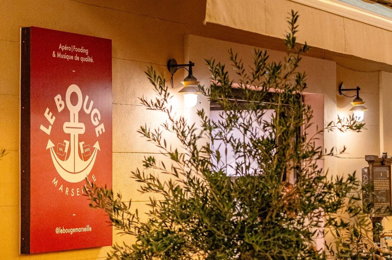 Enseigne rouge d'un établissement avec un logo d'ancre, éclairée par des lampes murales, partiellement masquée par un…