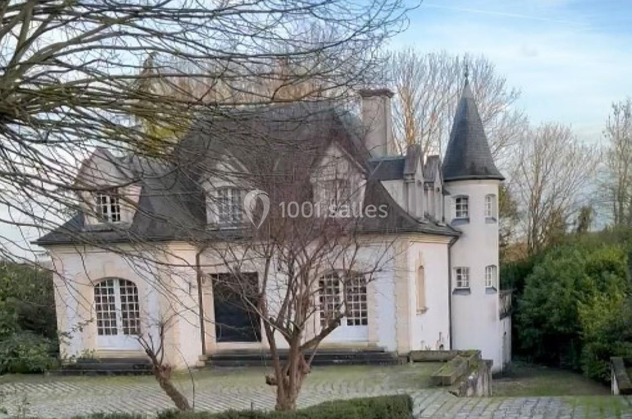 Grande maison blanche avec tourelle, entourée d'arbres et d'une allée pavée, sous un ciel dégagé.