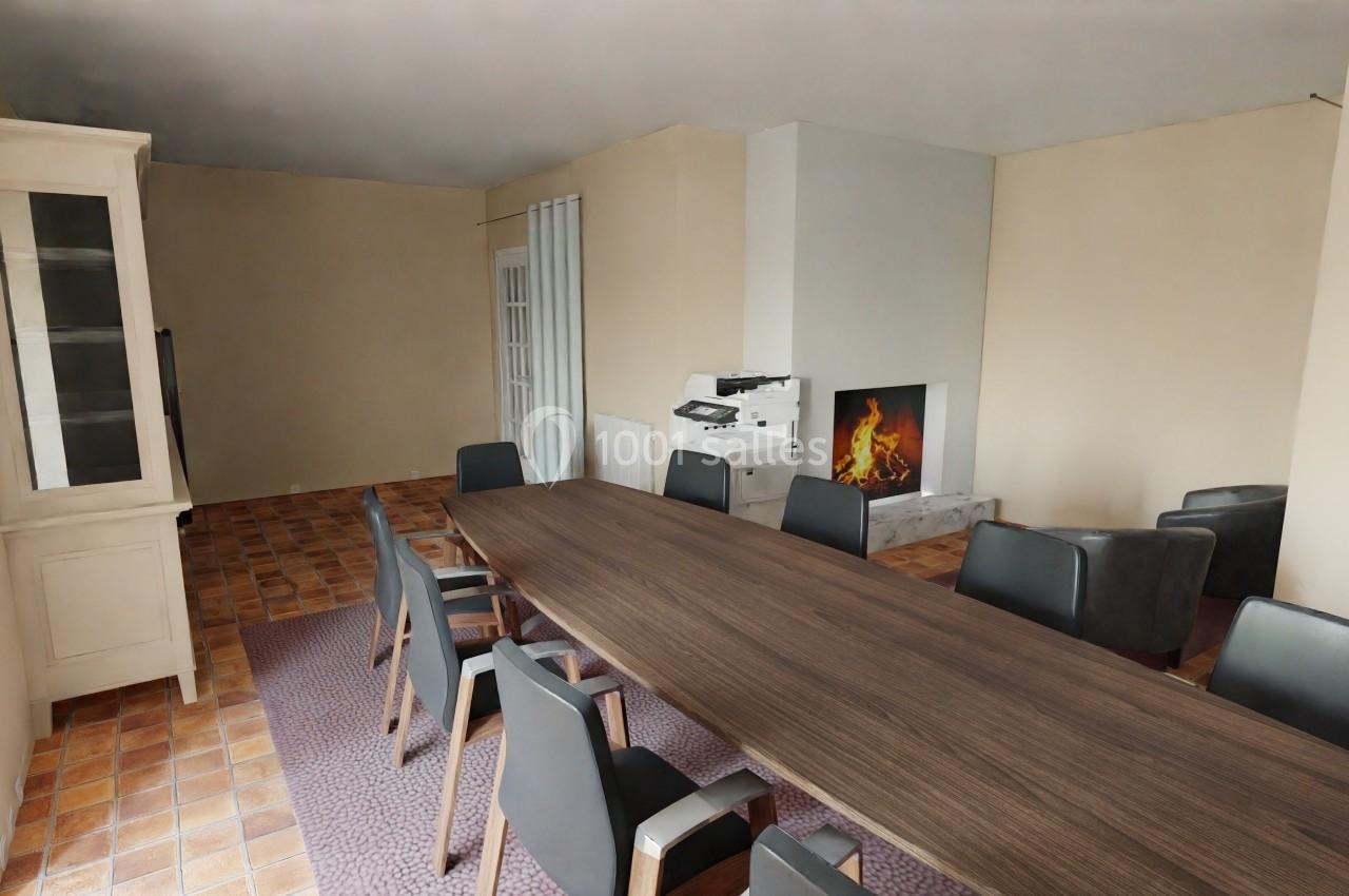 Salle de réunion avec une grande table en bois, chaises noires, cheminée allumée et sol en carrelage.