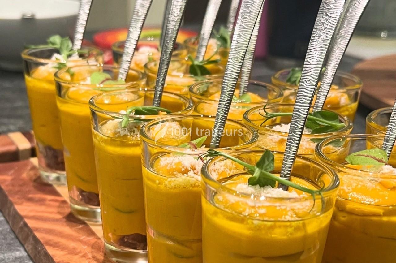Verrines de velouté orange garnies de feuilles de basilic et de cuillères en métal, présentées sur un plateau en bois.
