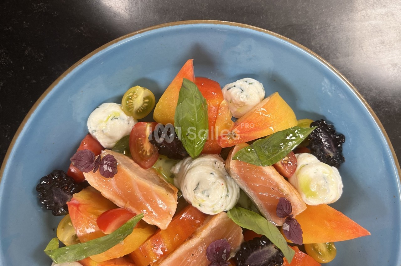 Assiette colorée de saumon fumé, tomates variées, mûres, herbes fraîches et quenelles de fromage sur une assiette bleue.
