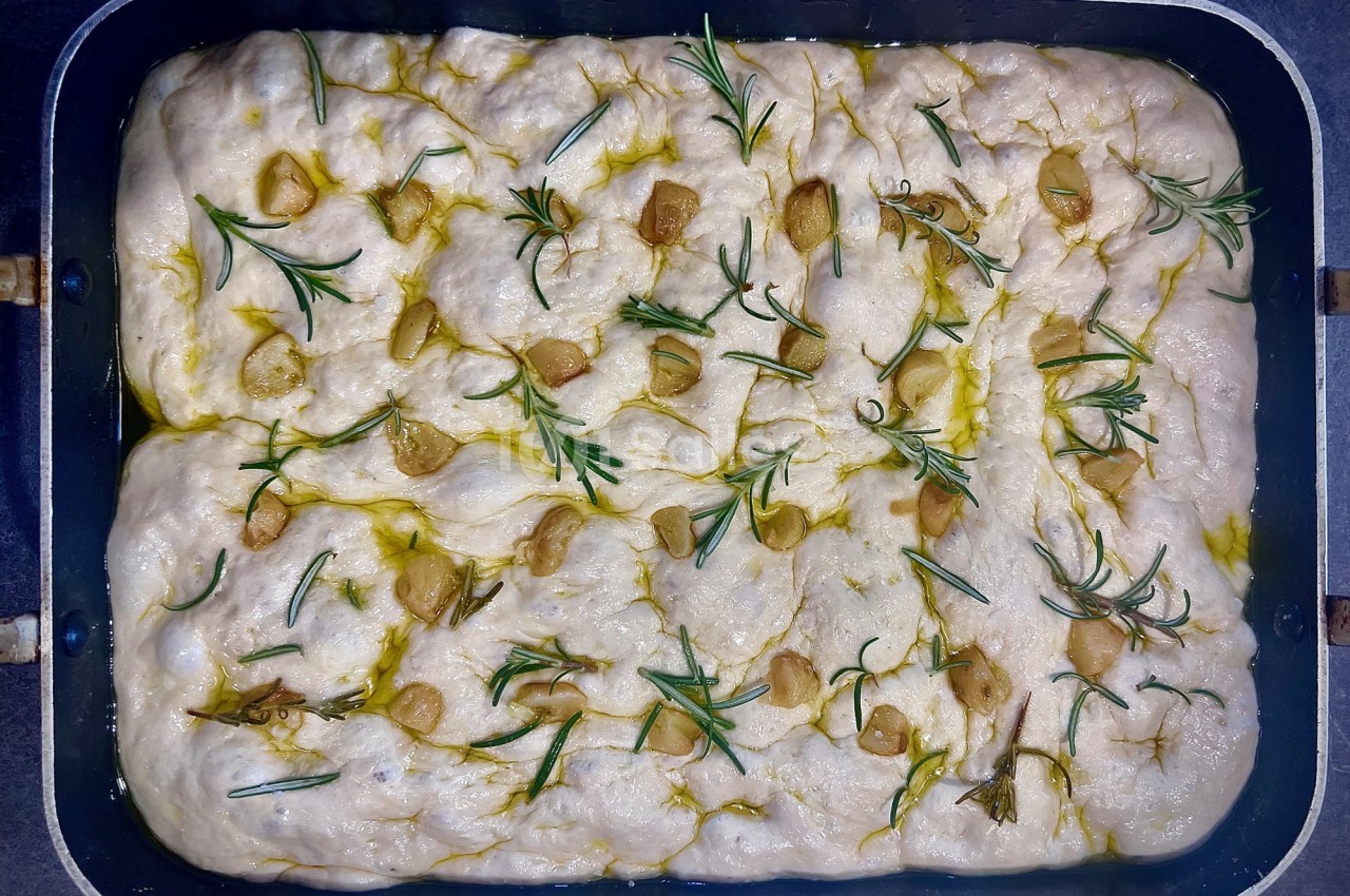 Pâte à focaccia crue garnie de romarin, d'ail et d'huile d'olive dans un moule rectangulaire.