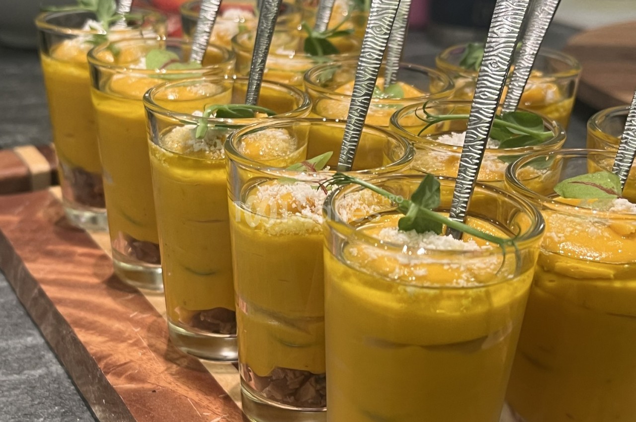 Verrines de velouté orange garnies de feuilles vertes, parmesan râpé et cuillères argentées.