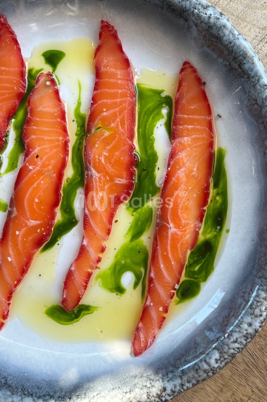 Tranches de saumon cru marinées, disposées avec de l'huile et une sauce verte sur une assiette en céramique.
